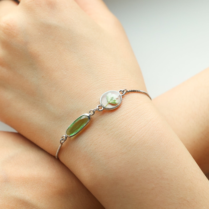 May include: A silver bracelet with a green oval-shaped gemstone and a round clear charm with a pressed flower inside.