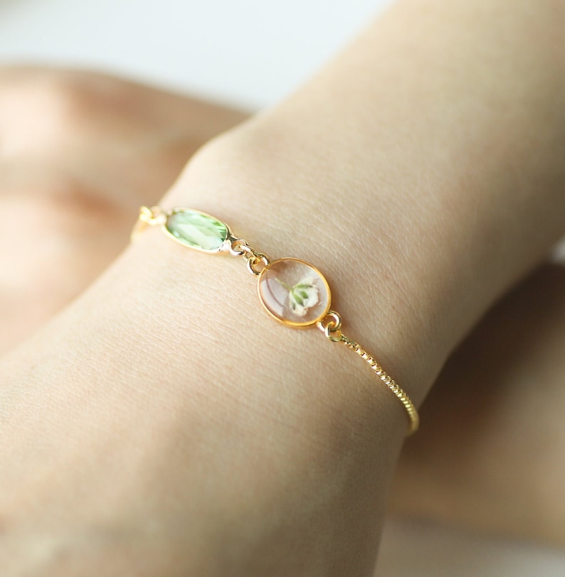 May include: A delicate gold chain bracelet with a small oval-shaped pendant featuring a pressed white flower. The bracelet also has a green oval-shaped gemstone.