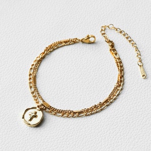 May include: A gold chain bracelet with a layered design and a small cross pendant. The pendant is white with a gold cross.