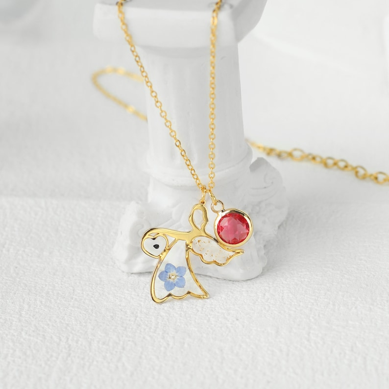 May include: Gold necklace with a delicate angel pendant featuring a blue flower and a red gemstone. The angel is holding a heart in its hand.