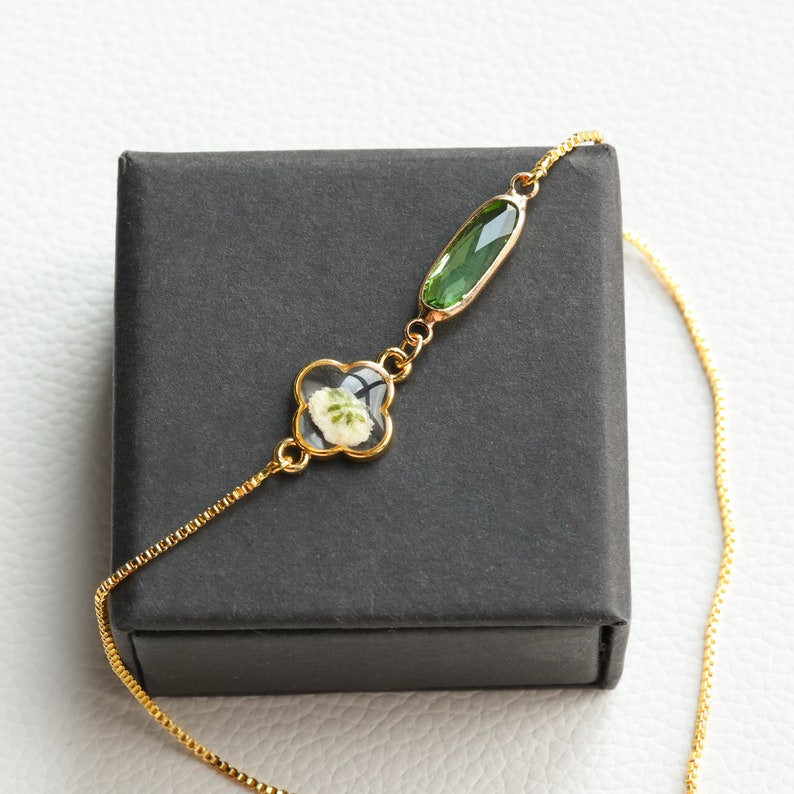 May include: A gold chain necklace with a green gemstone and a four-leaf clover charm with a pressed flower inside.