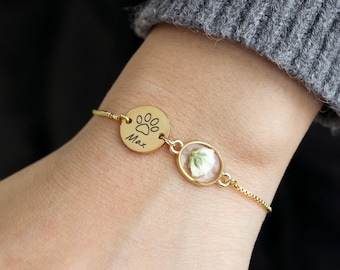 Puede incluir: Pulsera dorada con dos colgantes: uno redondo con una huella de pata y el nombre "Max", y otro ovalado con una flor blanca. La pulsera se lleva en la muñeca, con un jersey gris en el fondo.