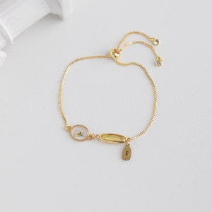 Puede incluir: Un brazalete ajustable de oro con un peque&ntilde;o dije ovalado que contiene una flor prensada y un dije rectangular m&aacute;s grande con una piedra amarilla. El brazalete tiene una peque&ntilde;a etiqueta con la palabra "Leo" grabada.