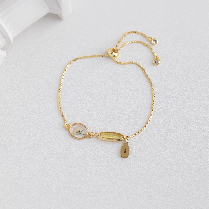 Puede incluir: Un brazalete ajustable de oro con un peque&ntilde;o dije ovalado que contiene una flor prensada y un dije rectangular m&aacute;s grande con una piedra amarilla. El brazalete tiene una peque&ntilde;a etiqueta con la palabra "Leo" grabada.