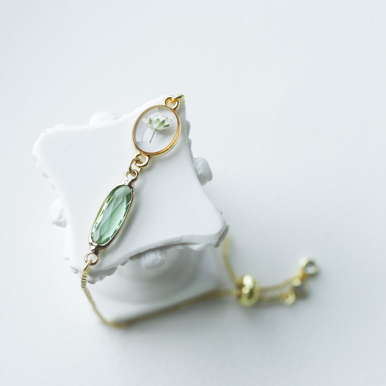 May include: A gold chain bracelet with a green oval-shaped gemstone and a round pendant with a dried flower inside.