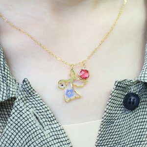 May include: A gold necklace with a delicate chain and a pendant featuring a small angel with pressed flowers and a red gemstone.