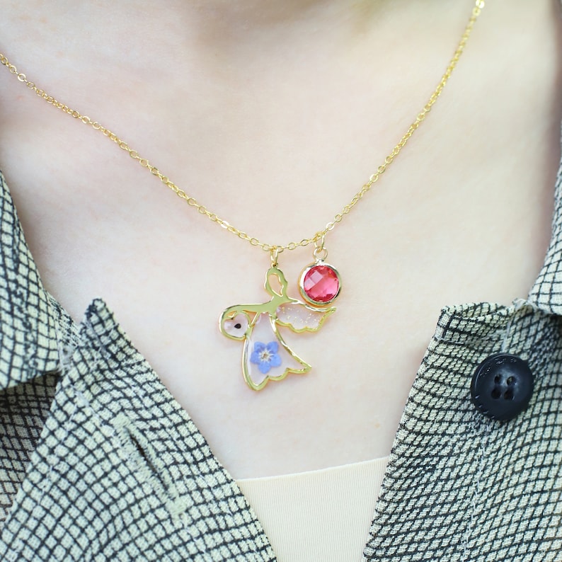 May include: A gold necklace with a delicate chain and a pendant featuring a small angel with pressed flowers and a red gemstone.