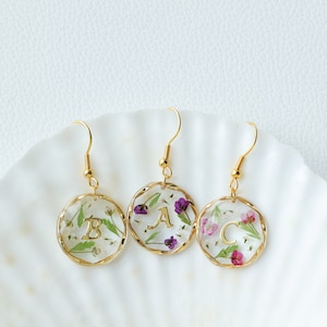 May include: Three gold earrings with a round, clear resin design. Each earring features a different letter, A, B, and C, pressed into the resin. The resin is filled with small, dried flowers in shades of purple and white.