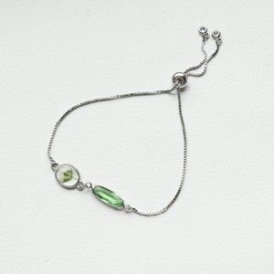 May include: A silver adjustable bracelet with a green oval-shaped gemstone and a clear oval-shaped pendant with a pressed flower inside.