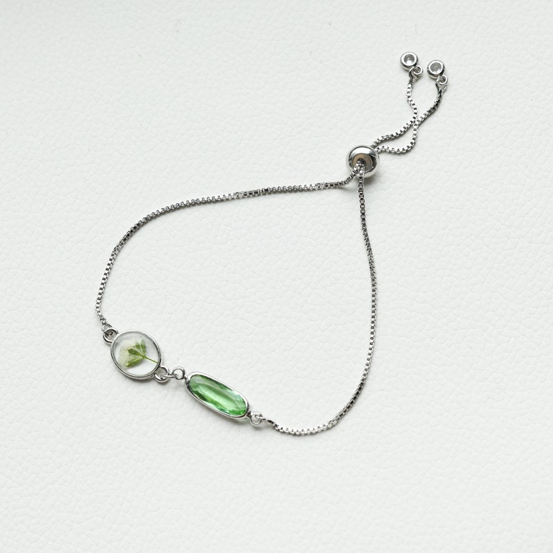 May include: A silver adjustable bracelet with a green oval-shaped gemstone and a clear oval-shaped pendant with a pressed flower inside.