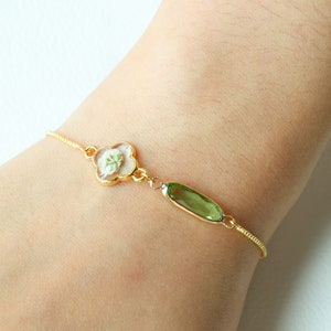 May include: A gold chain bracelet with a clover-shaped charm containing a pressed white flower and a green oval charm.
