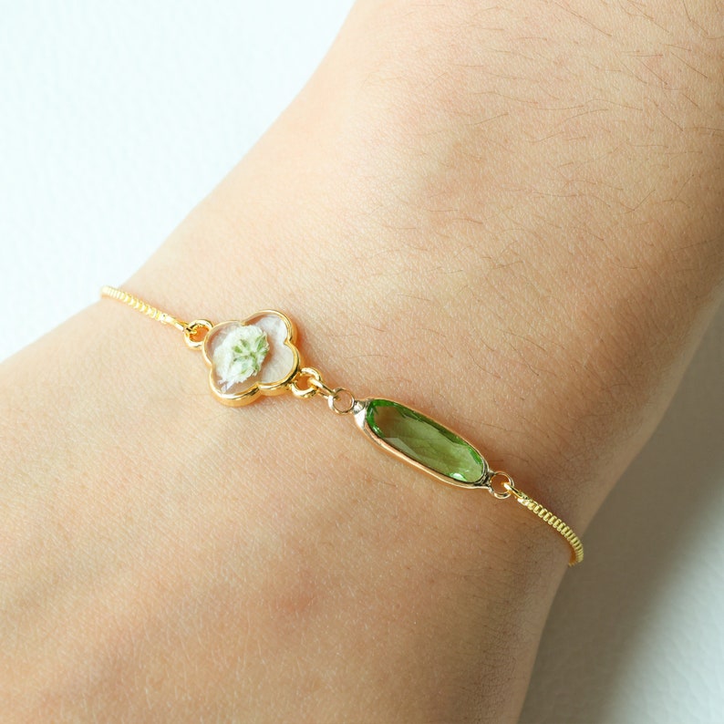 May include: A gold chain bracelet with a clover-shaped charm containing a pressed white flower and a green oval charm.