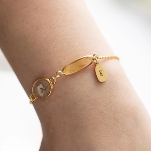 May include: A gold-toned chain bracelet featuring a circular pendant containing a pressed white flower, an elongated oval amber-colored charm, and a small rectangular gold-toned tag engraved with the name Leo in black script lettering.