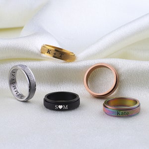 May include: Five different engraved rings with various designs and colors. The rings are made of metal and have a brushed finish. The engravings include initials, roman numerals, and a heart symbol.
