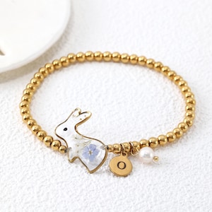 May include: A gold-toned beaded bracelet featuring a clear resin charm shaped like a rabbit with embedded dried flowers. The bracelet includes a small circular gold-toned charm engraved with the letter O and a single white pearl-like bead accent.