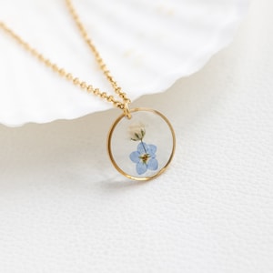 May include: A gold chain necklace with a round pendant featuring a pressed blue forget-me-not flower.