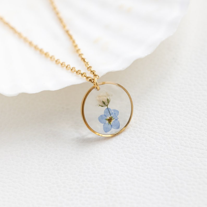 May include: A gold chain necklace with a round pendant featuring a pressed blue forget-me-not flower.