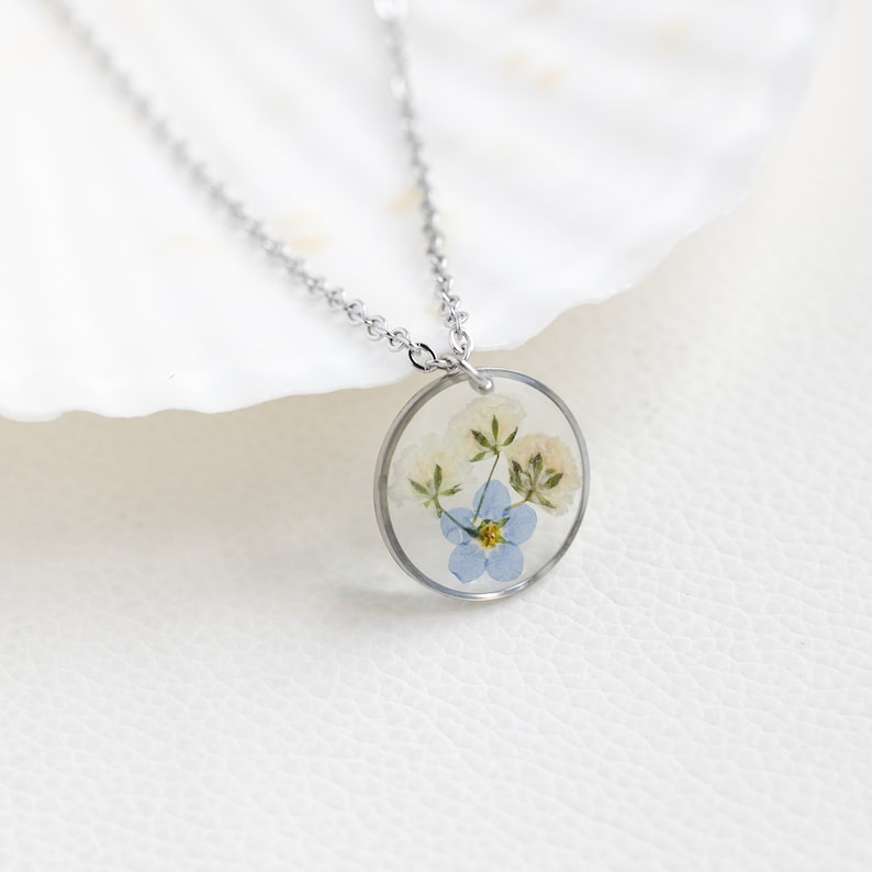 May include: A silver chain necklace with a round pendant featuring pressed blue and white flowers encased in clear resin.