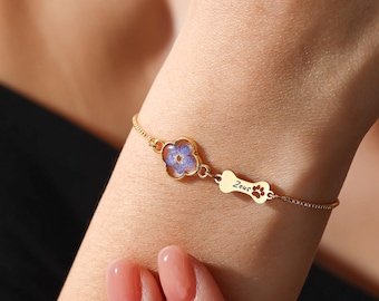 Puede incluir: Pulsera dorada con una flor de cuatro pétalos con diseño floral azul y un dije en forma de hueso con el nombre "Zeus" y huellas de patas. La pulsera es delicada y elegante.