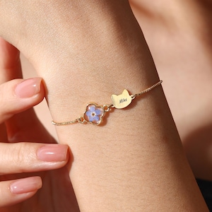May include: A gold bracelet with a small, four-petal flower with blue petals set in resin. A cat-shaped charm with the name "Miles" is attached. The bracelet is worn on a wrist.