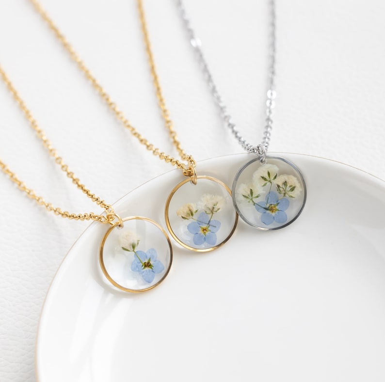 May include: Two delicate necklaces with round pendants featuring pressed forget-me-not flowers. One necklace has a gold chain and the other has a silver chain.