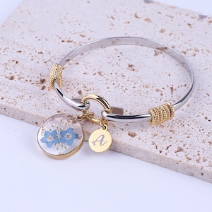 May include: A silver and gold bangle bracelet with a charm featuring a pressed flower design and a gold charm with the letter 'A'.