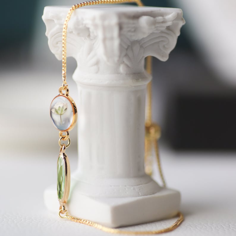 May include: A gold chain bracelet with a delicate white flower encased in resin and a green glass bead. The bracelet is displayed on a white pedestal.