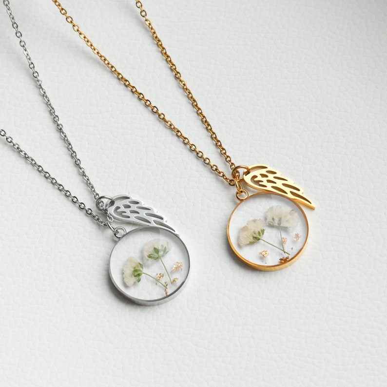 K&ouml;nnte beinhalten: Zwei zarte Halsketten mit goldenen und silbernen Ketten. Jede Halskette hat einen runden Anh&auml;nger mit einem gepressten Blumenmuster und einem kleinen Fl&uuml;gel-Anh&auml;nger. Die Anh&auml;nger sind klar und zeigen die zarten Details der Blumen.