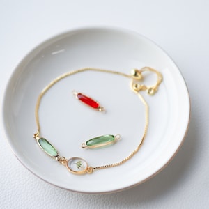 May include: A gold chain bracelet with a green and clear glass oval charm, a green glass rectangle charm, and a red glass rectangle charm. The bracelet is on a white plate.