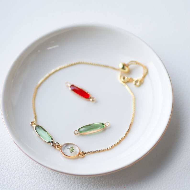 May include: A gold chain bracelet with a green and clear glass oval charm, a green glass rectangle charm, and a red glass rectangle charm. The bracelet is on a white plate.