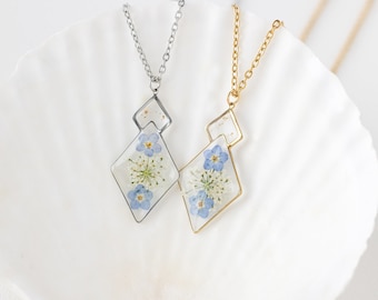 May include: Two necklaces with diamond-shaped pendants, each containing blue and white dried flowers encased in clear resin. One necklace has a silver chain and frame, the other a gold chain and frame. The pendants hang on a white surface.