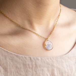 May include: A gold chain necklace with a round pendant featuring pressed flowers in shades of blue and white.