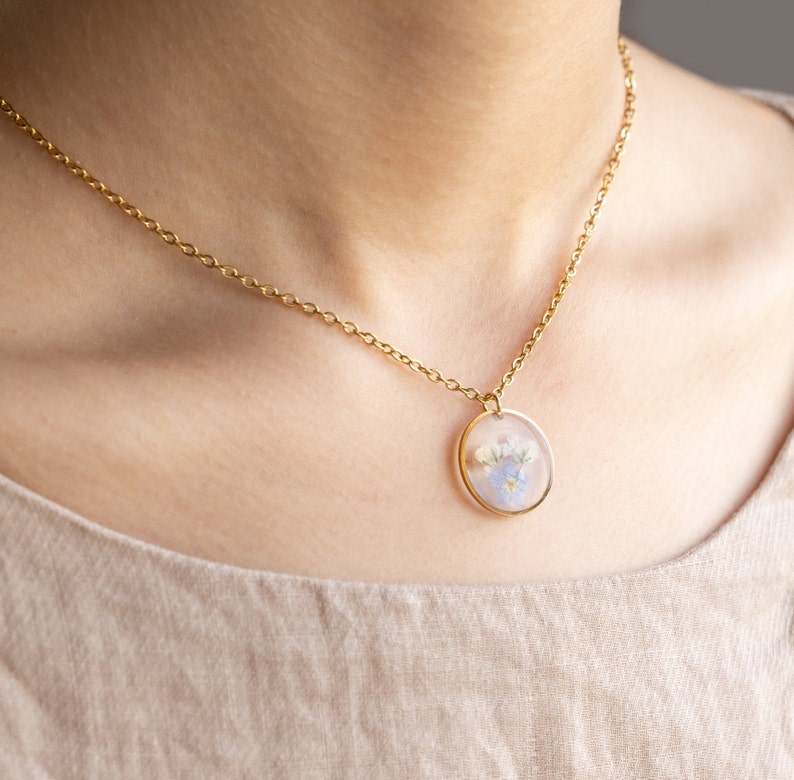 May include: A gold chain necklace with a round pendant featuring pressed flowers in shades of blue and white.