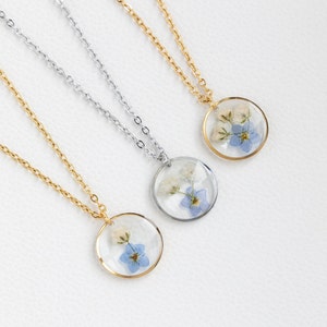 May include: Three delicate necklaces with round pendants featuring pressed forget-me-not flowers and white blossoms encased in clear resin. The necklaces are available in gold, silver, and a combination of both.