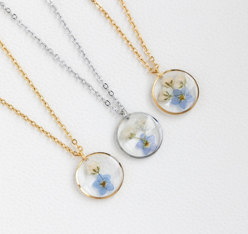 May include: Three delicate necklaces with round pendants featuring pressed forget-me-not flowers and white blossoms encased in clear resin. The necklaces are available in gold, silver, and a combination of both.
