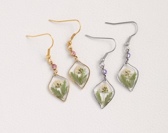 May include: A pair of dangle earrings with a delicate flower pressed inside a clear resin teardrop shape. One pair has gold-tone hooks and pink crystals, the other has silver-tone hooks and purple crystals.