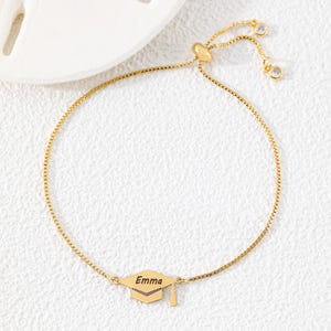 Op de afbeelding: Een goudkleurige verstelbare armband met een afstudeerhoed-bedel met de naam "Emma" gegraveerd. De armband heeft een fijne ketting en twee kleine, heldere kristallen accenten bij de sluiting.