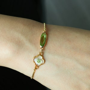 May include: A gold bracelet with a green gemstone and a white flower encased in resin. The bracelet is on a person's wrist.