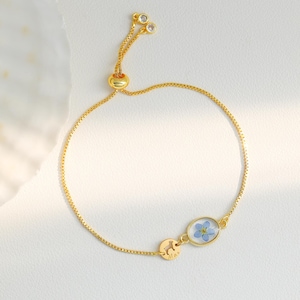 May include: A gold chain bracelet with a small oval charm featuring a pressed blue flower. The bracelet has a sliding clasp and two small clear stones at the top.