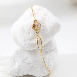 Puede incluir: Una pulsera dorada con un dije de trébol de cuatro hojas con un patrón marrón claro y una piedra transparente de forma ovalada. La pulsera se muestra sobre una superficie blanca texturizada.