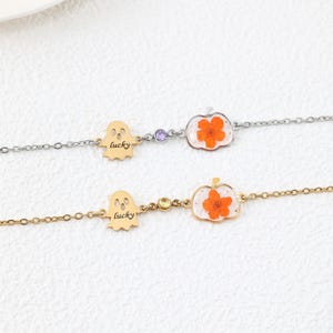 May include: Two delicate bracelets, one silver-toned and one gold-toned, each featuring a ghost-shaped charm with the word lucky engraved, a small round gemstone, and a pumpkin-shaped pendant containing a pressed orange flower.