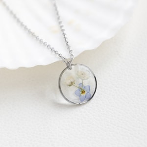 May include: A silver chain necklace with a round pendant featuring pressed white and blue flowers encased in clear resin.