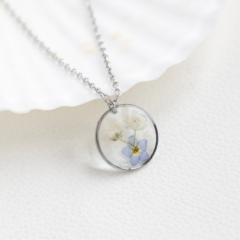 May include: A silver chain necklace with a round pendant featuring pressed white and blue flowers encased in clear resin.