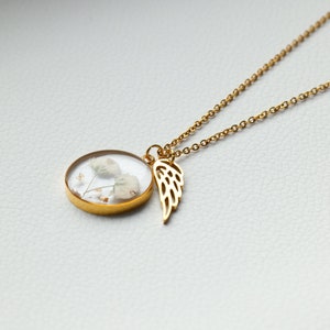 K&ouml;nnte beinhalten: Goldkette mit einem runden Anh&auml;nger, der getrocknete wei&szlig;e Blumen und einen goldenen Engelsfl&uuml;gel-Anh&auml;nger enth&auml;lt.