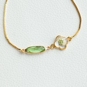 May include: A gold chain bracelet with a green oval-shaped gemstone and a gold clover charm with a pressed white flower inside.