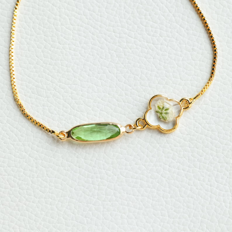 May include: A gold chain bracelet with a green oval-shaped gemstone and a gold clover charm with a pressed white flower inside.