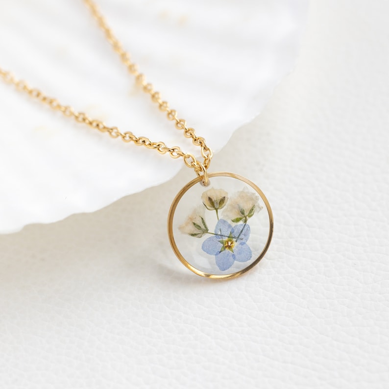 May include: A gold chain necklace with a round pendant featuring pressed forget-me-not flowers in shades of blue and white.