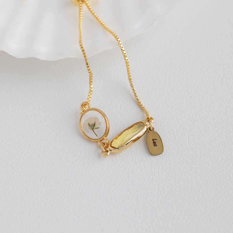 Puede incluir: Un brazalete de cadena dorada con un peque&ntilde;o colgante redondo que contiene una flor prensada y un colgante rectangular con la palabra "Leo" grabada.
