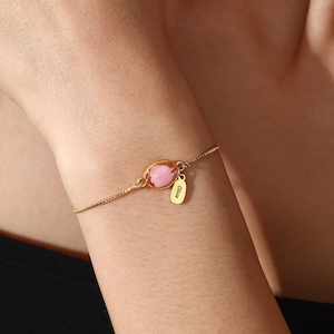 Puede incluir: Una pulsera dorada con una piedra preciosa rosa de forma ovalada y una pequeña etiqueta grabada con el nombre "Olivia". La pulsera es de diseño delicado, con una fina cadena. La piedra está engastada en un bisel dorado.