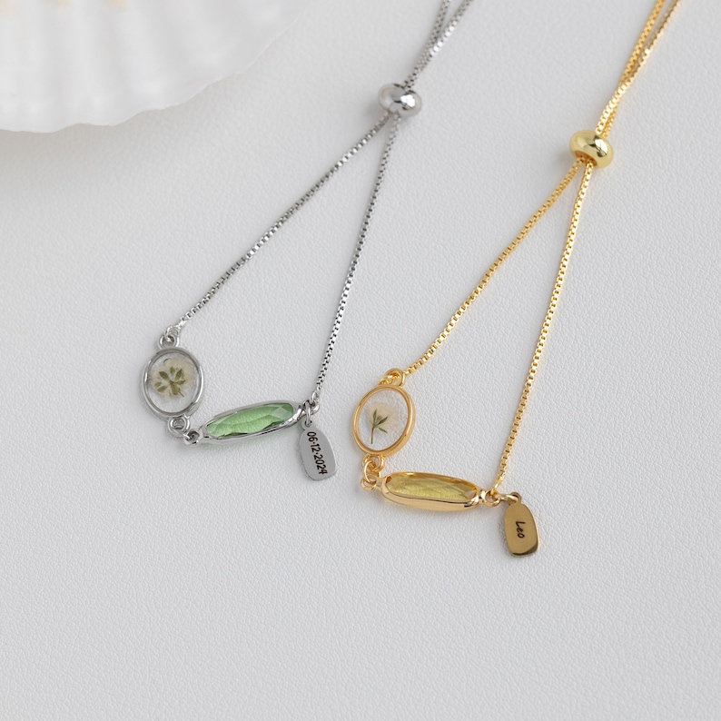 Puede incluir: Dos pulseras ajustables con cadenas delicadas, cada una con un peque&ntilde;o dije ovalado con una flor prensada en su interior. Una pulsera tiene una cadena plateada y un dije verde con la fecha "06.12.2014" grabada en una etiqueta. La otra pulsera tiene una cadena dorada y un dije amarillo con la palabra "Leo" grabada en una etiqueta.